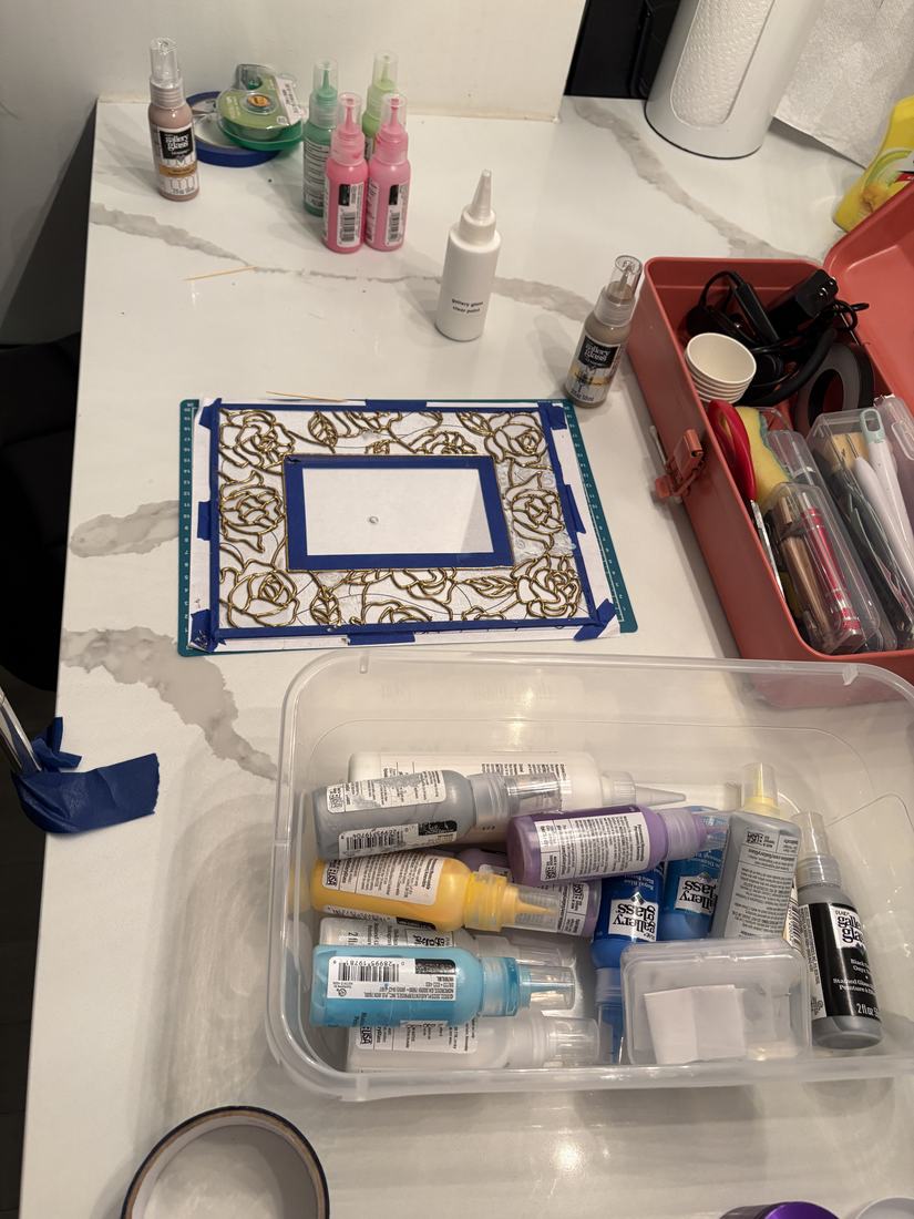 A small crafting station covered in stained glass paint bottles and a rose-patterned glass piece in progress.