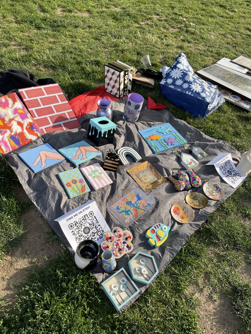 A blanket spread out on grass in a park, covered with handmade art and crafts for sale, with Find Me on Etsy QR code signs.
